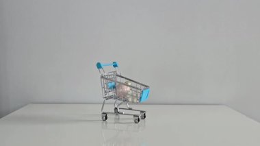 Gri arka planı olan beyaz bir masada mini oyuncak alışveriş arabası. El beyaz ve mavi Noel süsleri ekliyor, neşeli cazibe için kırmızı bir takı ile bitiriyor..