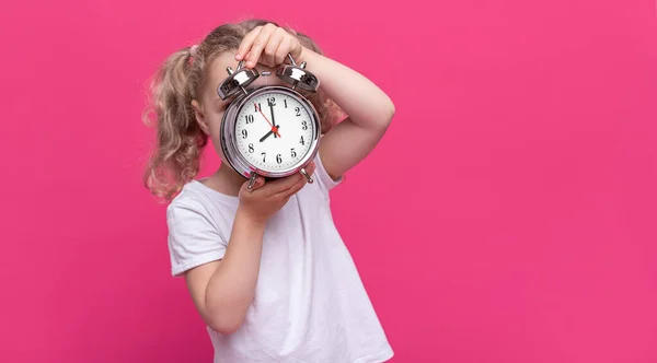 小さな女の子がピンクの背景に大きな目覚まし時計を持っています — ストック写真