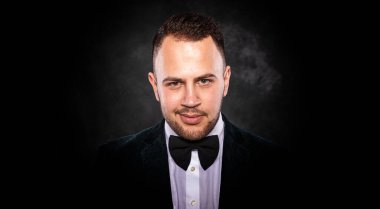 Sakallı, özgüvenli bir genç adam karanlık bir arka plana poz veriyor..