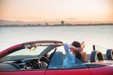 Woman in red car with open roof at background of sea water. Travel, freedom and holidays concept.
