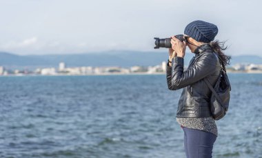 Genç bir fotoğrafçı kadın bir deniz manzarasının arka planında fotoğraf ya da video çekiyor..