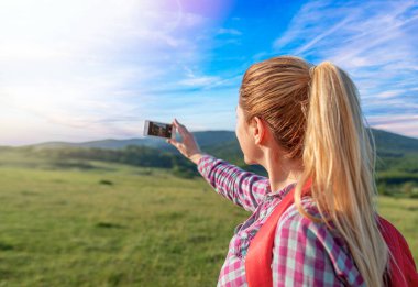 A traveler girl photographs a natural landscape on her phone.