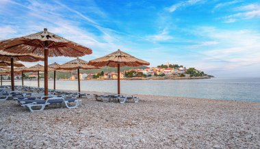 Sahilde güneşli yataklı saman şemsiyeler. Gün batımında ya da gün doğumunda kumsalda saman şemsiyeler ve güverte sandalyeleri. Primosten, Hırvatistan.