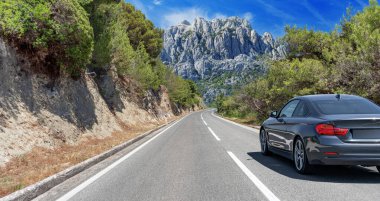 Araba, güneşli bir günde, kayalık dağların arka planına doğru yol alır..