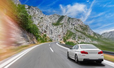 Güneşli bir günde, beyaz bir araba kayalık dağların arka planına doğru yol alır..