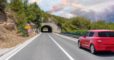 Kırmızı bir araba, güneşli bir günde, kayalık dağların ve tünellerin arka planına doğru yol alır..