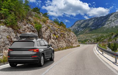 Yolculuk arabası, güneşli bir günde, kayalık dağların arka planına karşı otoyol boyunca gider..