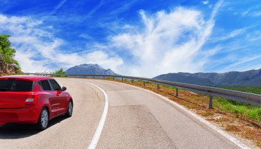 Kırmızı bir araba, güneşli bir günde, kayalık dağların ve tünellerin arka planına doğru yol alır..