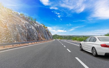 Güneşli bir günde, beyaz bir araba kayalık dağların arka planına doğru yol alır..