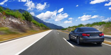 Gri bir araba güneşli bir günde otoyol boyunca kayalık dağların arka planına doğru gidiyor..