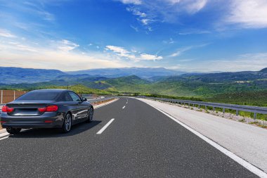 Gri bir araba güneşli bir günde otoyol boyunca kayalık dağların arka planına doğru gidiyor..