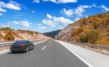 Güneşli bir günde, siyah bir araba kayalık dağların arka planına doğru yol alır..