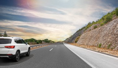 Güneşli bir günde, beyaz bir araba kayalık dağların arka planına doğru yol alır..