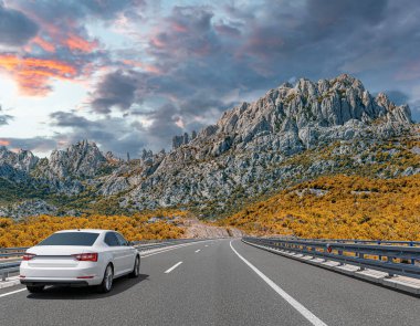 Güneşli bir günde, beyaz bir araba kayalık dağların arka planına doğru yol alır..
