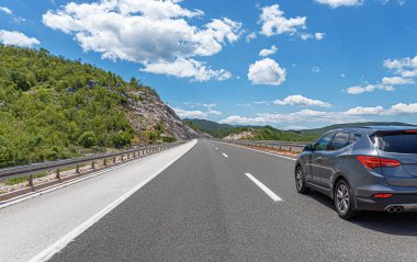 Gri bir araba güneşli bir günde otoyol boyunca kayalık dağların arka planına doğru gidiyor..