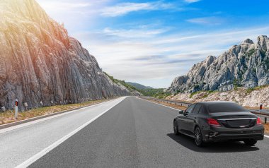 Gri bir araba güneşli bir günde otoyol boyunca kayalık dağların arka planına doğru gidiyor..