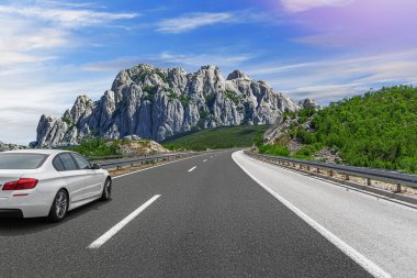 Güneşli bir günde, beyaz bir araba kayalık dağların arka planına doğru yol alır..