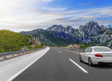 Güneşli bir günde, beyaz bir araba kayalık dağların arka planına doğru yol alır..