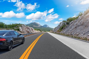 Güneşli bir günde, siyah bir araba kayalık dağların arka planına doğru yol alır..