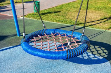 Modern bir oyun parkında kırmızı ağlı mavi yuvarlak bir salıncak askıya alınır. Bölge, yeşillik ve yollarla çevrili, güvenlik için kauçuk bir yüzeye sahip..