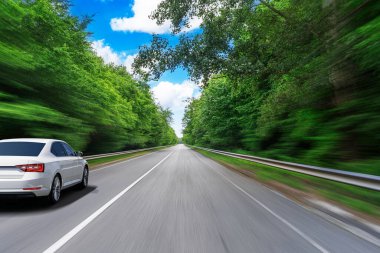 Parlak mavi gökyüzünün altında yemyeşil ağaçlarla çevrili pürüzsüz bir otoyolda, parlak, beyaz bir sedan yüksek hızda sürüyor, dinamik bir hareket ve özgürlük hissi yaratıyor..