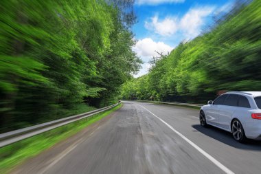 Parlak mavi gökyüzünün altında, yoğun yeşil ormanlarla çevrili, dolambaçlı bir otoyolda, hareketin ve maceranın önemini vurgulayan hareketli, bulanık bir etki yaratan parlak beyaz bir araba hızlanır..