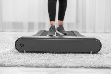 Siyah tayt ve gri spor ayakkabı giyen bir kişi modern, minimalist bir ev ortamında küçük bir koşu bandında yürüyor, fitness ve aktif bir yaşam tarzına odaklanıyor..