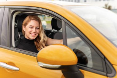 Gülümseyen sarışın bir kadın sarı bir arabanın şoför koltuğunda oturur, sıcak ve kendinden emin bir ifadeyle kameraya bakar..