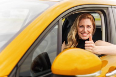 Kendine güvenen sarışın bir kadın sarı bir arabada oturuyor, gülümsüyor ve pencereden baş parmak işareti yapıyor, başarıyı, mutluluğu ve bağımsızlığı simgeliyor..