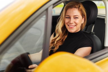 Gülümseyen sarışın bir kadın sarı bir arabanın şoför koltuğunda oturur, sıcak ve kendinden emin bir ifadeyle kameraya bakar..