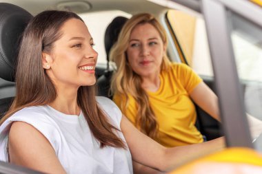 İki kadın sarı bir arabada oturur, keyifli bir anı paylaşırken şoför kendinden emin bir şekilde gülümserken arkadaşı hayranlıkla bakar, arkadaşlığı ve macerayı simgeler..