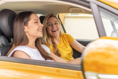 İki neşeli kadın sarı bir arabanın içinde eğlenceli ve hayat dolu bir anın tadını çıkarıyorlar. Birlikte yaptıkları yolculukta gülüyorlar ve keyifli bir deneyimi paylaşıyorlar..