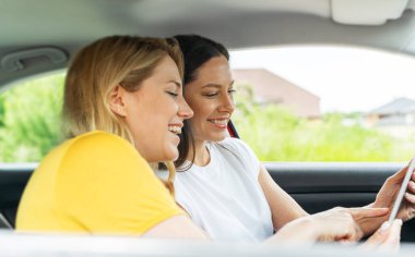 Arabada oturmuş gülümseyen iki kadın bir tablet kullanıyor. Bir yolculuk sırasında seyrüsefer ya da planlama yapıyor gibi görünüyorlar..