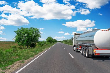 Büyük bir yakıt tankeri otoban boyunca ilerliyor. Etrafı yemyeşil yemyeşil ve açık mavi gökyüzü ile çevrili. Dağınık beyaz bulutlar, huzurlu kırsal alanı yansıtıyor..