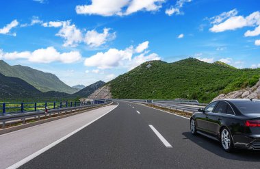 Parlak, mavi bir gökyüzünün altında, bulutlarla dolu yemyeşil dağları saran, bakımlı bir otoyol boyunca parlak, siyah bir sedan sürüyordu..