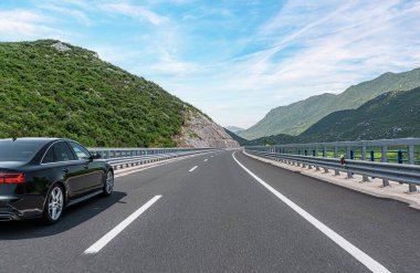 Parlak, mavi bir gökyüzünün altında, bulutlarla dolu yemyeşil dağları saran, bakımlı bir otoyol boyunca parlak, siyah bir sedan sürüyordu..