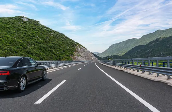 Parlak, mavi bir gökyüzünün altında, bulutlarla dolu yemyeşil dağları saran, bakımlı bir otoyol boyunca parlak, siyah bir sedan sürüyordu..