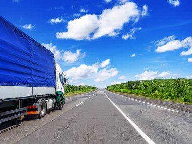 Mavi muşamba kaplı karavanı olan büyük bir kargo kamyonu, açık mavi gökyüzü altında ve dağınık bulutlar altında yemyeşil bir manzarayla çevrili düz bir otoyolda seyahat ediyor.