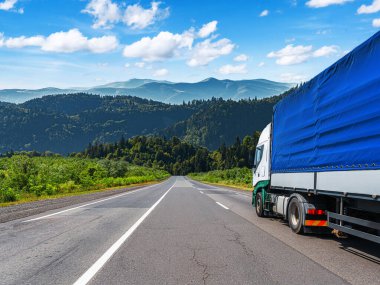 Muşamba kaplı kargo alanına sahip büyük bir kamyon yeşil tepeler ve parlak mavi bir gökyüzünün altındaki dağlarla çevrili manzaralı bir otoyol boyunca ilerliyor..