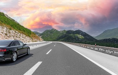 Siyah lüks bir sedan, turuncu, pembe ve mor renkli çarpıcı bir günbatımı gökyüzünün altında, yemyeşil dağlardan geçen dolambaçlı bir otoyol boyunca ilerliyor..