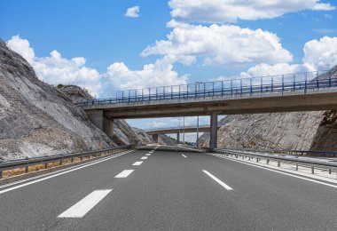 Modern, boş bir otoyol, dağlık bir arazide uzanır. Birden fazla beton köprünün altından geçer. Parlak mavi gökyüzü altında, dağınık bulutlarla birlikte..