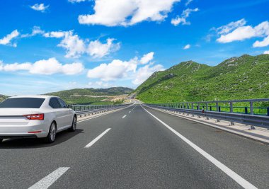 Beyaz bir sedan, yemyeşil dağlarla çevrili modern bir otoyol boyunca, dağınık bulutlu parlak mavi bir gökyüzünün altında, manzaralı ve huzurlu bir yolculuk atmosferi yaratır..