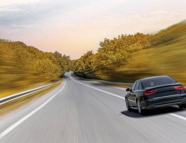 Siyah lüks bir sedan, sonbahar renklerinde ağaçlarla çevrili kıvrımlı bir otoyol boyunca hızla ilerliyor. Hareket bulanıklığı, hızlı hareketini vurguluyor..