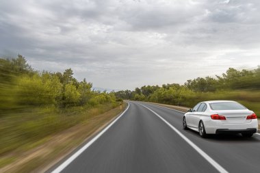 Beyaz bir sedan, bulutlu bir gökyüzünün altında, kıvrımlı, ağaçlarla kaplı bir otoyolda hızla ilerliyor. Manzaralı bir kırsal bölgede hızlı bir yolculuğun özünü yakalıyor..