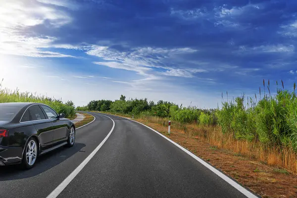 Siyah bir sedan kavisli, ağaçlarla kaplı bir otoyolun kenarına park edilmiş. Parlak mavi bir gökyüzünün altında, dağınık bulutlarla, manzaralı bir kırsal yolculukta huzurlu bir anı vurguluyor..