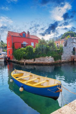 Yat limanında demirlemiş mavi ve sarı bir kayığın bulunduğu büyüleyici bir sahil manzarası. Etrafı tarihi taş mimarisi, canlı kırmızı bir ev ve parlak mavi gökyüzü altında yemyeşil bir yer..