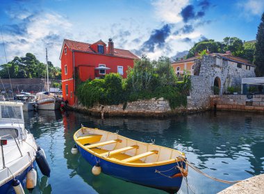 Yat limanında demirlemiş mavi ve sarı bir kayığın bulunduğu büyüleyici bir sahil manzarası. Etrafı tarihi taş mimarisi, canlı kırmızı bir ev ve parlak mavi gökyüzü altında yemyeşil bir yer..