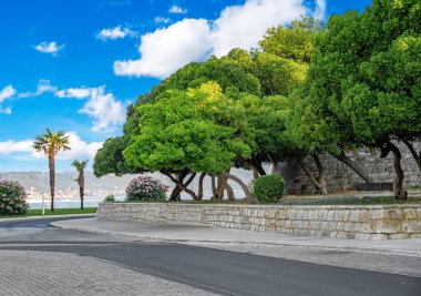 Bereketli yeşil ağaçları, palmiyeleri ve taş bir duvarı olan pitoresk bir şehir parkı. Parlak mavi bir gökyüzünün altında manzaralı bir deniz manzarası üzerine kurulmuş..