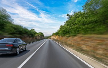 Parlak siyah bir araba ağaçlarla kaplı bir otoyolda hızla ilerliyor. Güneşli bir günde hız ve özgürlük hissini vurgulayan hareket bulanıklığı etkisi yaratıyor..