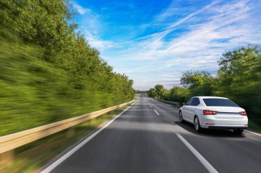 Beyaz bir sedan, parlak mavi bir gökyüzünün altında hızla yol alır. Hareket bulanıklığı, yolculuğun hızını ve özgürlüğünü yakalar..